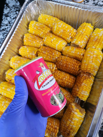 Corn on the cob in a foil pan with a can of Zdeco seasoning held by a gloved hand.